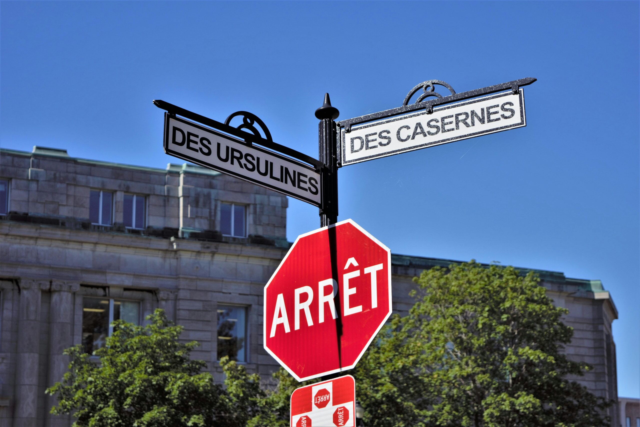 Bilingual street signs with stop sign against a clear blue sky in a city setting, featuring historical street names.