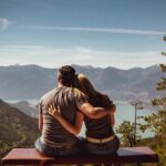 Couple sitting on bench embracing scenic mountain view, embodying romantic nature escape.