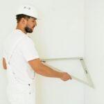 Side view of a construction worker with safety helmet measuring a wall corner indoors.