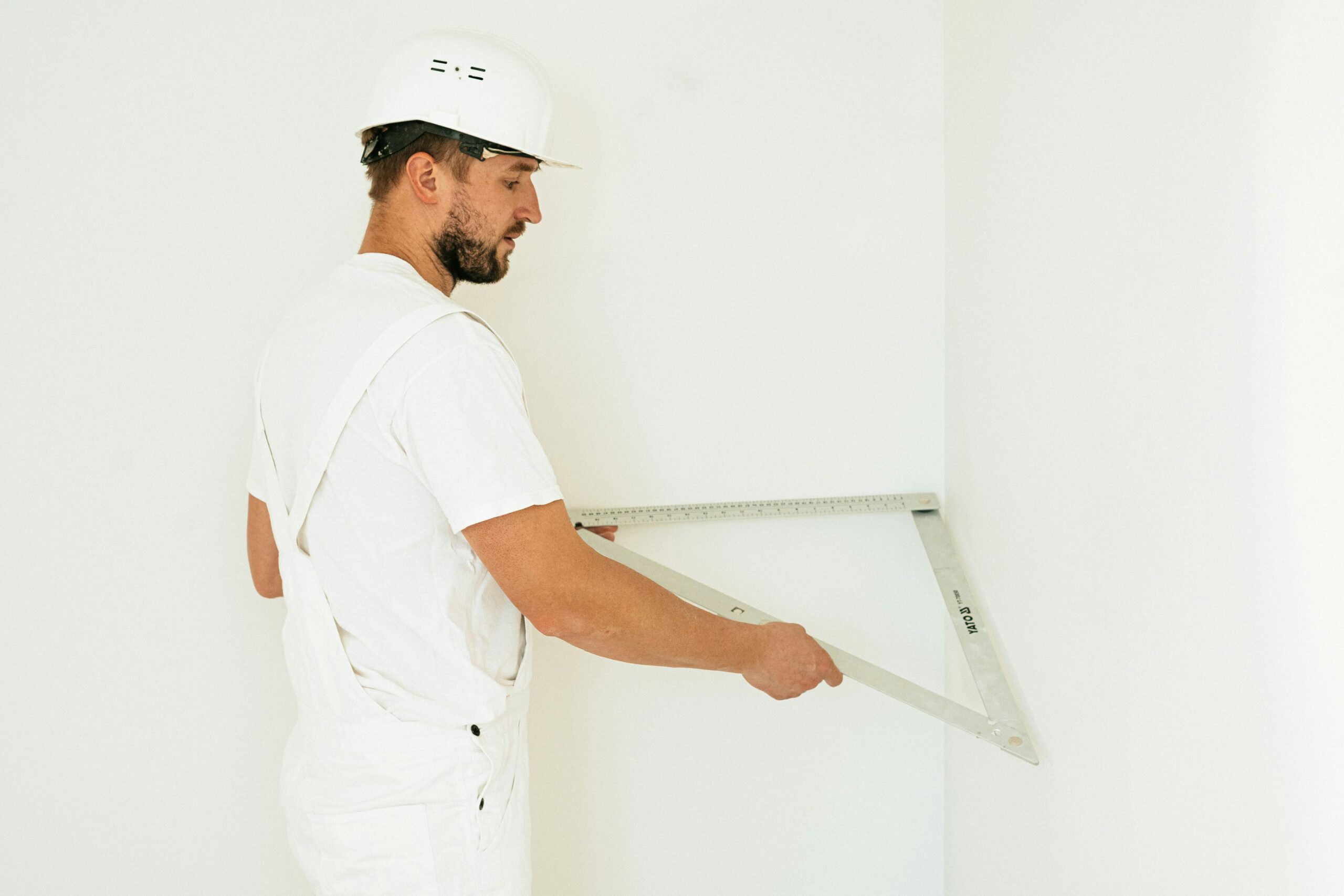 Side view of a construction worker with safety helmet measuring a wall corner indoors.